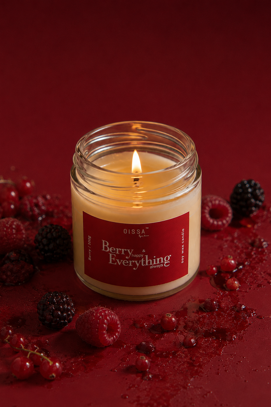 A lit glass jar candle with a red label reading 'Berry Everything' sits on a deep red surface surrounded by fresh berries and berry juice splatter. The brand name 'Qissa by su.i' is visible on the label.