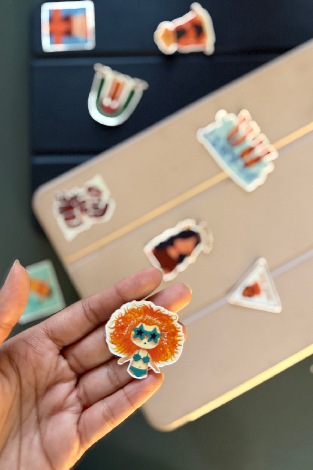 Hand holding a colorful sticker with a character design, with more stickers on a surface in the background.